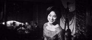Movie still from “When a Woman Ascends the Stairs” (1960), directed by Mikio Naruse – A woman in a dress smiling for the camera; Medium shot, Low angle