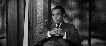 Movie still from “When a Woman Ascends the Stairs” (1960), directed by Mikio Naruse – A man in a suit sitting in front of a wooden wall; Medium shot, Low angle