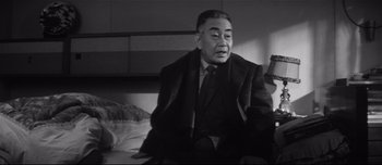 Movie still from “When a Woman Ascends the Stairs” (1960), directed by Mikio Naruse – An older man sitting on top of a bed in a room; Medium shot, Low angle