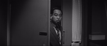 Movie still from “When a Woman Ascends the Stairs” (1960), directed by Mikio Naruse – A man in a suit and tie standing next to a door; Medium shot, Low angle
