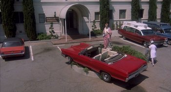 Movie still from “Where the Buffalo Roam” (1980), directed by Art Linson – A woman standing in front of a red car; Wide shot, High angle