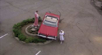 Movie still from “Where the Buffalo Roam” (1980), directed by Art Linson – A red car parked in the middle of a parking lot next to a statue; Extreme Wide shot, High angle