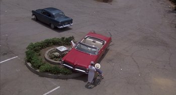 Movie still from “Where the Buffalo Roam” (1980), directed by Art Linson – A man standing next to a red car in a parking lot; Extreme Wide shot, High angle