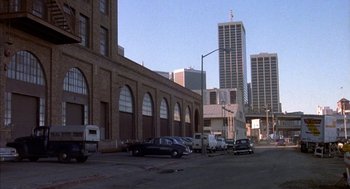 Movie still from “Where the Buffalo Roam” (1980), directed by Art Linson – Cars are parked on the side of the road near a tall building; Extreme Wide shot, High angle