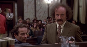 Movie still from “Where the Buffalo Roam” (1980), directed by Art Linson – A man in a suit and tie in front of a group of people in a court room; Medium shot, Low angle