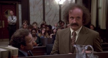 Movie still from “Where the Buffalo Roam” (1980), directed by Art Linson – A man in a suit and tie in front of a group of people in a court room; Medium shot, Low angle