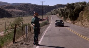 Movie still from “Where the Buffalo Roam” (1980), directed by Art Linson – A man standing on the side of a road next to a car; Wide shot, Over the shoulder angle