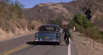 Movie still from “Where the Buffalo Roam” (1980), directed by Art Linson – A man riding a bike next to an old car; Wide shot, Over the shoulder angle