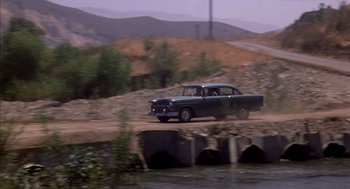 Movie still from “Where the Buffalo Roam” (1980), directed by Art Linson – An old car driving down a dirt road near a bridge; Wide shot, High angle