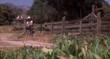 Movie still from “Where the Buffalo Roam” (1980), directed by Art Linson – A man riding a horse down a dirt road next to a wooden fence; Wide shot, High angle