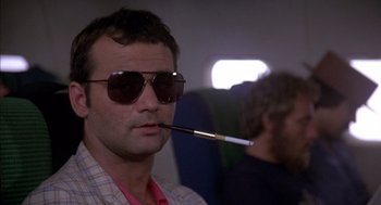 Movie still from “Where the Buffalo Roam” (1980), directed by Art Linson – A man wearing sunglasses and holding a cigarette in his mouth while sitting on an airplane; Close Up shot, Low angle