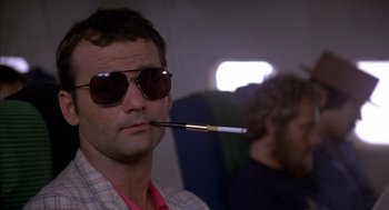 Movie still from “Where the Buffalo Roam” (1980), directed by Art Linson – A man wearing sunglasses and holding a cigarette in his mouth while sitting on a plane; Close Up shot, Low angle