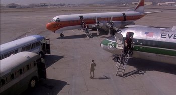 Movie still from “Where the Buffalo Roam” (1980), directed by Art Linson – An airplane parked on the tarmac with people boarding it; Extreme Wide shot, High angle