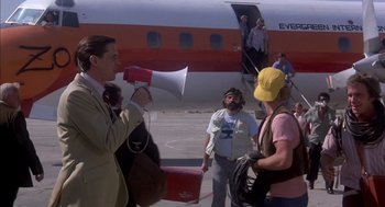 Movie still from “Where the Buffalo Roam” (1980), directed by Art Linson – A group of people standing on a tarmac near an airplane; Wide shot, Over the shoulder angle