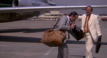 Movie still from “Where the Buffalo Roam” (1980), directed by Art Linson – A man carrying luggage walking across an airport tarmac; Medium shot, Low angle