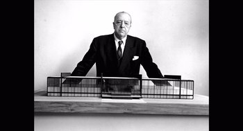 Movie still from “Where'd You Go, Bernadette” (2019), directed by Richard Linklater – An older man in a suit and tie standing in front of a building model; Medium shot, Low angle