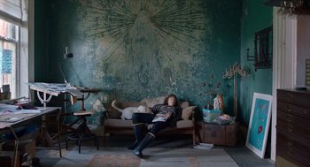 Movie still from “Where'd You Go, Bernadette” (2019), directed by Richard Linklater – A woman sitting on top of a couch in front of a painting; Wide shot, Low angle