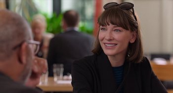 Movie still from “Where'd You Go, Bernadette” (2019), directed by Richard Linklater – A woman sitting at a table in front of a group of people; Close Up shot, Over the shoulder angle