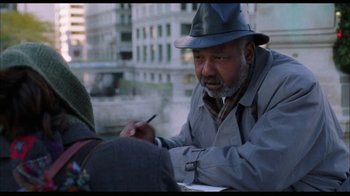 Movie still from “While You Were Sleeping” (1995), directed by Jon Turteltaub – An older man wearing a hat is writing on a piece of paper; Medium shot, Over the shoulder angle