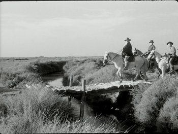 Movie still from “White Mane” (1953), directed by Albert Lamorisse – Two men riding horses across a bridge over a river; Wide shot, Low angle