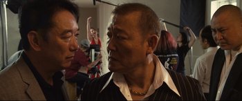Movie still from “Why Don't You Play in Hell?” (2013), directed by Sion Sono – A man in a black suit and a white shirt; Close Up shot, Over the shoulder angle