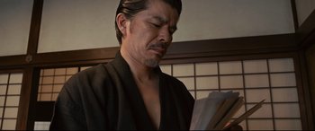 Movie still from “Why Don't You Play in Hell?” (2013), directed by Sion Sono – A man wearing a black robe reading a book; Close Up shot, Low angle