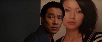 Movie still from “Why Don't You Play in Hell?” (2013), directed by Sion Sono – A man standing in front of a picture of a woman; Close Up shot, Over the shoulder angle