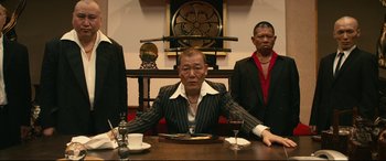 Movie still from “Why Don't You Play in Hell?” (2013), directed by Sion Sono – A man sitting at a table with a plate of food; Medium shot, Low angle
