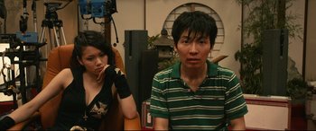 Movie still from “Why Don't You Play in Hell?” (2013), directed by Sion Sono – A man and a woman sitting next to each other in a room; Medium shot, High angle