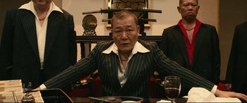 Movie still from “Why Don't You Play in Hell?” (2013), directed by Sion Sono – An older man sitting at a table with a sword on top of his head; Medium shot, Low angle