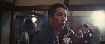 Movie still from “Why Don't You Play in Hell?” (2013), directed by Sion Sono – A man in a black shirt is in front of a group of people; Medium shot, Low angle