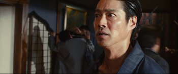 Movie still from “Why Don't You Play in Hell?” (2013), directed by Sion Sono – A person in a room with a painting on the wall; Close Up shot, Low angle