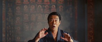 Movie still from “Why Don't You Play in Hell?” (2013), directed by Sion Sono – An asian man in a black robe making a funny face in front of a wall of buddha images; Medium shot, Low angle