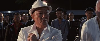 Movie still from “Why Don't You Play in Hell?” (2013), directed by Sion Sono – An older man wearing a white suit and a white hat; Close Up shot, Over the shoulder angle