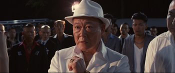 Movie still from “Why Don't You Play in Hell?” (2013), directed by Sion Sono – An older man wearing a white hat and a white suit; Close Up shot, Over the shoulder angle