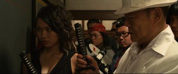 Movie still from “Why Don't You Play in Hell?” (2013), directed by Sion Sono – A group of people standing in a room; Close Up shot, Over the shoulder angle