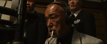 Movie still from “Why Don't You Play in Hell?” (2013), directed by Sion Sono – An older man wearing headphones while smoking a cigarette; Close Up shot, Low angle