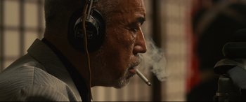 Movie still from “Why Don't You Play in Hell?” (2013), directed by Sion Sono – An older man wearing headphones and smoking a cigarette; Close Up shot, Low angle