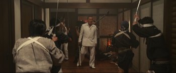 Movie still from “Why Don't You Play in Hell?” (2013), directed by Sion Sono – A man in a white suit is standing in front of a group of people; Wide shot, Low angle