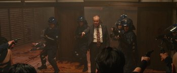 Movie still from “Why Don't You Play in Hell?” (2013), directed by Sion Sono – A group of people in a room with guns pointed at a man; Medium shot, High angle