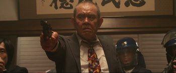 Movie still from “Why Don't You Play in Hell?” (2013), directed by Sion Sono – An older man wearing a suit and tie holding a gun in his right hand; Medium shot, Low angle