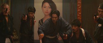Movie still from “Why Don't You Play in Hell?” (2013), directed by Sion Sono – A group of men standing in front of a poster of a woman; Medium shot, Low angle