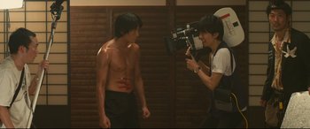 Movie still from “Why Don't You Play in Hell?” (2013), directed by Sion Sono – A man is being videotaped by another man; Medium shot, Low angle
