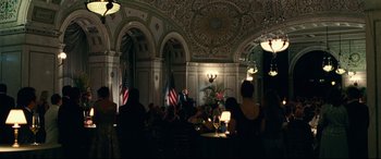Movie still from “Widows” (2018), directed by Steve McQueen – A group of people sitting at tables in front of a microphone; Wide shot, Over the shoulder angle