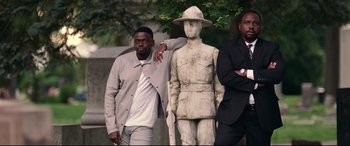 Movie still from “Widows” (2018), directed by Steve McQueen – A man standing next to a statue of an army man; Medium shot, Low angle