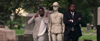 Movie still from “Widows” (2018), directed by Steve McQueen – A man standing next to a statue of a soldier; Medium shot, Low angle