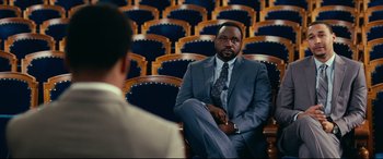 Movie still from “Widows” (2018), directed by Steve McQueen – A man in a suit sitting in front of another man in a suit; Medium shot, Over the shoulder angle