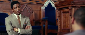 Movie still from “Widows” (2018), directed by Steve McQueen – A man sitting in a chair in front of a wooden chair; Medium shot, Low angle