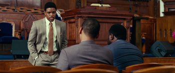 Movie still from “Widows” (2018), directed by Steve McQueen – A man in a suit is talking to a group of people; Medium shot, Over the shoulder angle