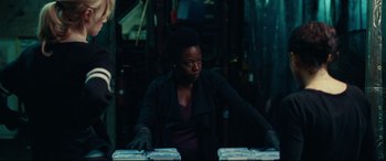 Movie still from “Widows” (2018), directed by Steve McQueen – A woman in black jacket and black gloves at a table; Medium shot, Low angle